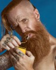 Thoughtful red haired bearded shirtless having tattoo on fingers and arms and with earrings in ear man with gray eyes and undercut hairstyle holding matchbox and lighting cigarette while frowning and looking at camera on isolated blue background — Stock Photo