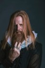 Frowned thoughtful severe red haired middle aged nobleman in clothes of Elizabethans era with white lush starchy ruff and tattoo on fingers touching beard and looking at camera on gray background — Stock Photo