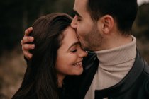 Vista laterale della coppia innamorata con gli occhi chiusi sorridenti mentre si abbracciano e si baciano lungo alberi di conifere durante il giorno con vento nuvoloso — Foto stock