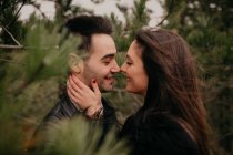 Vista lateral de la pareja enamorada con los ojos cerrados sonriendo mientras se abrazan y besan entre sí a lo largo de árboles de coníferas durante el día en tiempo nublado ventoso - foto de stock