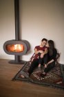 Happy couple in casual clothes embracing each other sitting on floor near fireplace on colorful carpet against white wall at home — Stock Photo