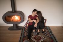 Casal feliz em roupas casuais abraçando uns aos outros sentado no chão perto da lareira no tapete colorido contra a parede branca em casa — Fotografia de Stock