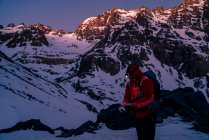 Человек в яркой куртке с рюкзаком и фонариком отдыхает, стоя в сумерках в окружении снежных горных вершин — стоковое фото