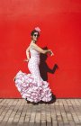 Vista lateral da mulher alegre excêntrica em traje rosa colorido sorrindo e dançando pelo fundo da parede vermelha no dia ensolarado — Fotografia de Stock