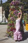 Vue latérale de femme excentrique curieuse en costume rose coloré debout près de beau mur de fleurs lumineuses le jour ensoleillé — Photo de stock