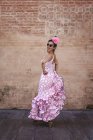 Боковой вид эксцентричной веселой женщины в красочном розовом костюме, улыбающейся и танцующей у кирпичной стены в солнечный день — стоковое фото