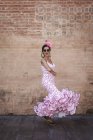 Vista lateral da mulher alegre excêntrica em traje rosa colorido sorrindo e dançando pela parede de tijolo no dia ensolarado — Fotografia de Stock