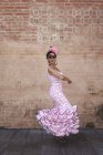 Vista lateral da mulher alegre excêntrica em traje rosa colorido sorrindo e dançando pela parede de tijolo no dia ensolarado — Fotografia de Stock