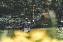 Visão traseira do homem equipado harling peixe enquanto em pé em waders em torrente de montanha por penhasco e floresta — Fotografia de Stock