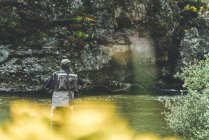 Back view of equipped man harling fish while standing in waders in mountain torrent by cliff and forest — Stock Photo