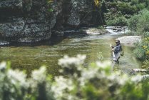 Вид сбоку на оборудованного человека, который рыбачит, стоя в болотах в горном потоке у скалы и леса — стоковое фото