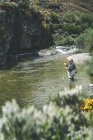 Вид сбоку на оборудованного человека, который рыбачит, стоя в болотах в горном потоке у скалы и леса — стоковое фото