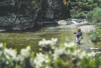 Side view of equipped man harling fish while standing in waders in mountain torrent by cliff and forest — Stock Photo