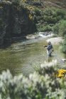 Vista lateral del hombre equipado harling pescado mientras está de pie en vadeadores en torrente de montaña por acantilado y bosque - foto de stock