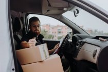 Corriere in bicchieri che prepara i pacchi per il trasporto mentre si siede e segna scatole in auto su sfondo sfocato durante il giorno — Foto stock