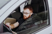 Человек фокусирует и проверяет документы, сидя за рулем в салоне автомобиля в дневное время на размытом фоне — стоковое фото