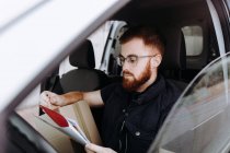 Человек фокусирует и проверяет документы, сидя за рулем в салоне автомобиля в дневное время на размытом фоне — стоковое фото