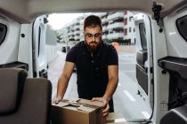 Courier in glasses carefully unloading cardboard boxes from car for further delivery to customer on blurred background during daytime — Stock Photo