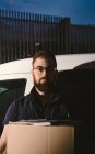 Дорослий бородатий чоловік в окулярах думає і дивиться на камеру, стоячи біля машини і тримаючи картонні коробки ввечері — стокове фото