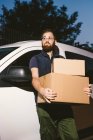 Hombre barbudo adulto en gafas pensando y mirando hacia otro lado mientras está de pie cerca del coche y sosteniendo cajas de cartón por la noche - foto de stock