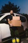 Взрослый бородатый мужчина в очках думает и смотрит в камеру, стоя возле машины и держа в руках картонные коробки вечером — стоковое фото