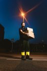Man in glasses carrying boxes while standing on street and looking at camera near glowing streetlight in evening on blurred background — Stock Photo