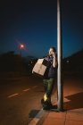 Mensageiro barbudo adulto em óculos com caixas fazendo chamada de advertência e olhando para longe enquanto de pé e apoiando-se no poste de luz de rua na rua à noite — Fotografia de Stock