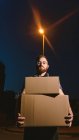 Homem de óculos carregando caixas enquanto em pé na rua e olhando para a câmera perto de luz de rua brilhante à noite no fundo borrado — Fotografia de Stock