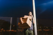 Courier in glasses with boxes making warning call to customer for further delivery and looking away while standing and leaning on streetlight post on street in evening — Stock Photo