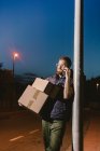 Courier in glasses with boxes making warning call and looking away while standing and leaning on streetlight post on street in evening — Stock Photo