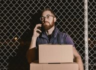 Courier in glasses with boxes looking at camera while standing and leaning on mesh fence talking on the mobile phone on street at night — Stock Photo