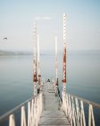 Узкий пирс с выветриваемыми перилами и дальний путешественник расположен вблизи спокойной морской воды против безоблачного неба — стоковое фото