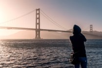 Back view of person in jacket with hood looking at long bridge under endless wavy sea in USA — Stock Photo