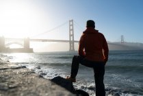 From behind man in warm clothes standing on coast by distant iron bridge over blue sea in sun rays in USA — Stock Photo