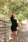 Pregnant woman looking at camera and touching belly while standing on road near garden in sunny day — Stock Photo