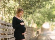 Pregnant woman looking away and touching belly while standing on road near garden in sunny day — Stock Photo