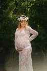 Femme enceinte avec les yeux fermés en robe de dentelle blanche touchant le ventre tout en se tenant dans la forêt — Photo de stock