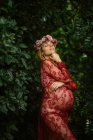 Mulher grávida com os olhos fechados tocando barriga e queixo enquanto em pé no jardim em dia ensolarado — Fotografia de Stock
