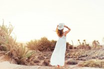 Frau in weißem Kleid berührt Hut auf trockenem Feld im Sonnenlicht — Stockfoto