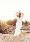 Жінка в білій сукні торкається капелюха в сухому полі на сонячному світлі — стокове фото