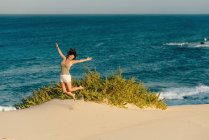 Активна жінка в білих шортах стрибає з розтягнутими руками на піщаному пляжі — стокове фото