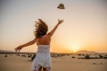 Rückenansicht einer schlanken Frau, die die Hände hebt und den Hut zum Sonnenuntergang im Wind in Fuerteventura, Las Palmas, Spanien mitwirft — Stockfoto