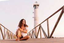 Nachdenklich gebräunte Frau im Sommerkleid sitzt mit überkreuzten Beinen auf Holzbrücke zum Leuchtturm in Fuerteventura, Las Palmas, Spanien — Stockfoto