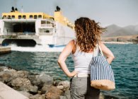 Rückenansicht einer aktiven Frau mit den Händen zur Seite, die auf ein großes Boot in türkisfarbenen Wellen am Pier in Fuerteventura, Las Palmas, Spanien blickt — Stockfoto