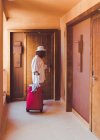 Frau in lässiger weißer Kleidung mit rotem Koffer schließt Holztür im Hotelflur — Stockfoto