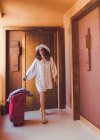 Mulher de óculos de sol e roupas brancas casuais com mala vermelha sorrindo e olhando para longe enquanto vai ao longo do corredor do hotel — Fotografia de Stock