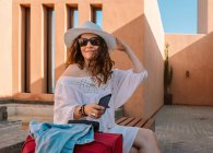 Happy woman in sunglasses and casual white dress looking away and touching hat while sitting on bench near building — Stock Photo
