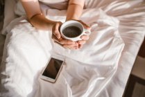 Von oben Hände einer Frau, die morgens eine Tasse Kaffee hält, während sie ihr Smartphone im Bett benutzt — Stockfoto