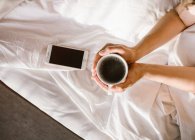 Зверху жінка тримає чашку кави, використовуючи смартфон в ліжку вранці — стокове фото