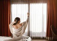 Вид сзади взрослой женщины с поднятой рукой, отводящей взгляд и касающейся волос, сидящей на кровати у закрытого окна — стоковое фото